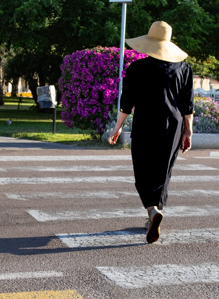 Una signora di spalle attraversa sulle strisce pedonali in una mattina d'estate, con un cappello di paglia. Sullo sfondo bouganville in fiore; sul prato a sinistra un piccione. A colori.